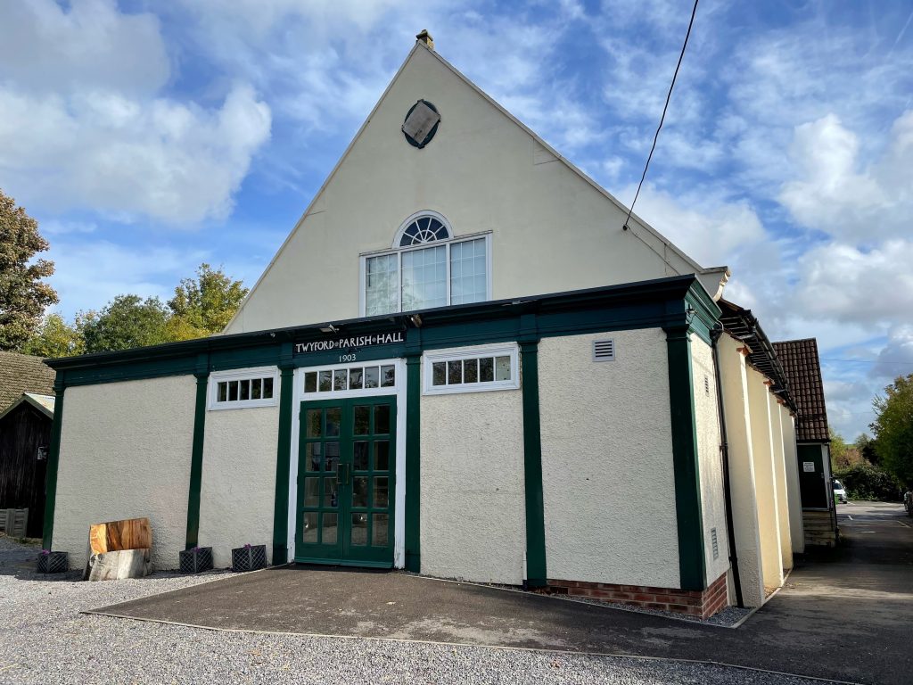 An outside view of Twyford Parish Hall. A single storey building with pointy roof, painted cream with dark green door and wood surrounds. 